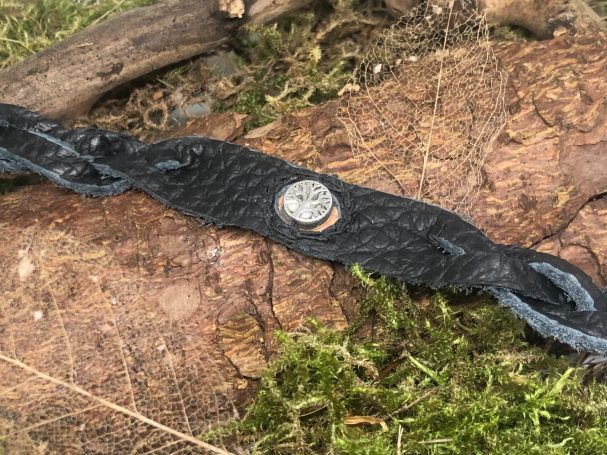 Lederarmband mit in der Mitte eingenähtem Charm, zu beiden Seiten sind geflochtene Enden, der Verschluss ist ein Mönchskopfverschluss